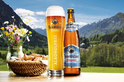 Ein Glas und eine Flasche Benediktiner Weissbier alkoholfrei stehen vor einer malerischen Berglandschaft, umgeben von einem Korb mit Brezeln und bunten Blumen.