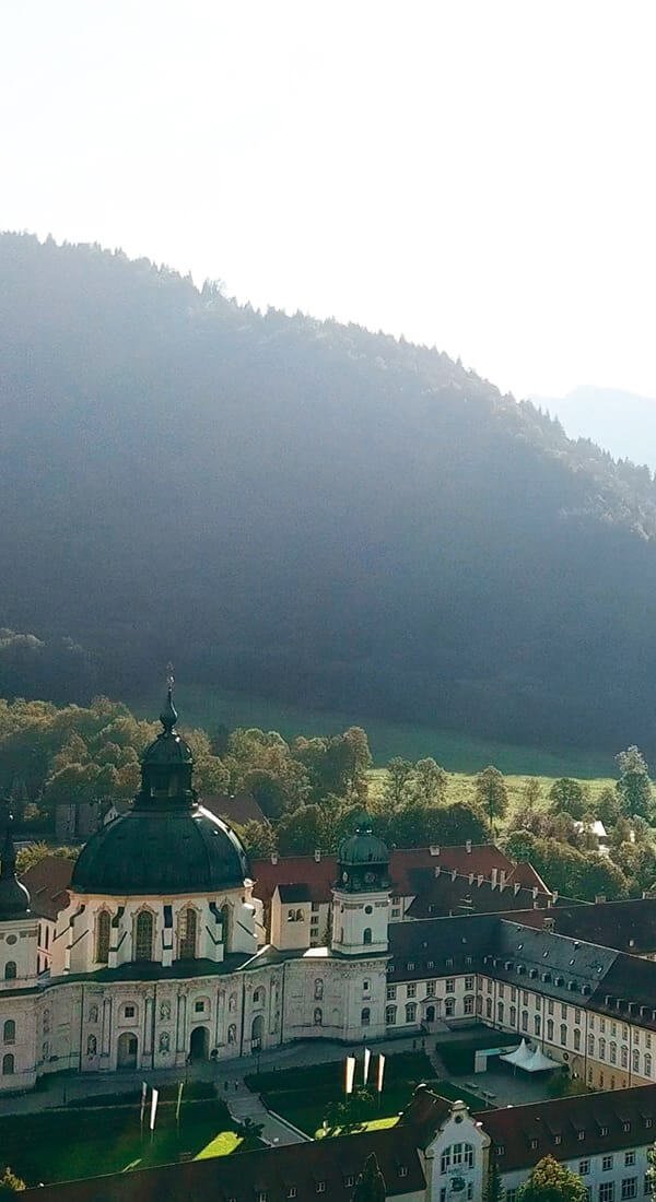 Blick auf das Kloster Ettal in den Ammergauer Alpen, umgeben von grünen Wiesen und Bergen im Hintergrund.